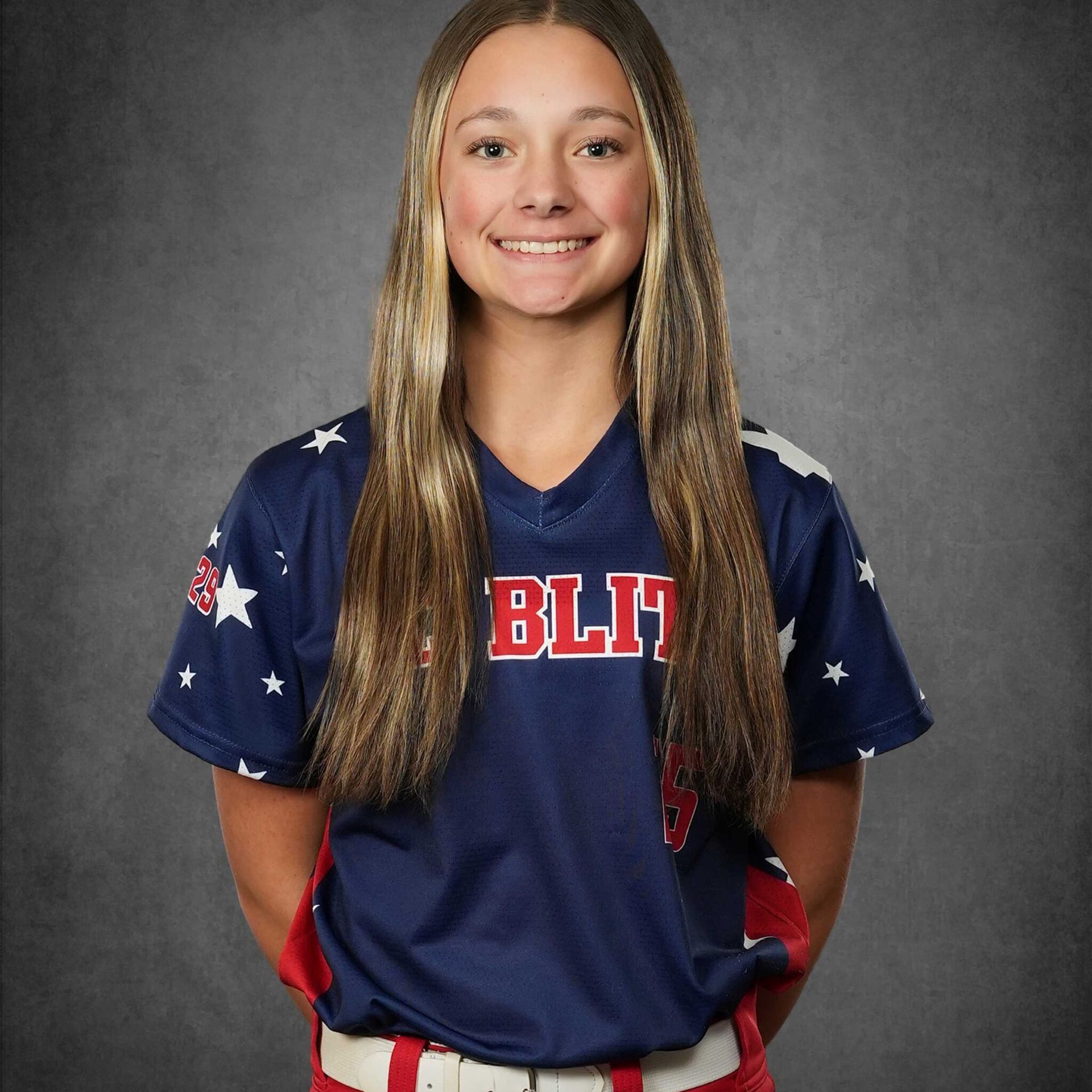 Young softball player in blue uniform with long blonde hair, smiling against a gray studio backdrop