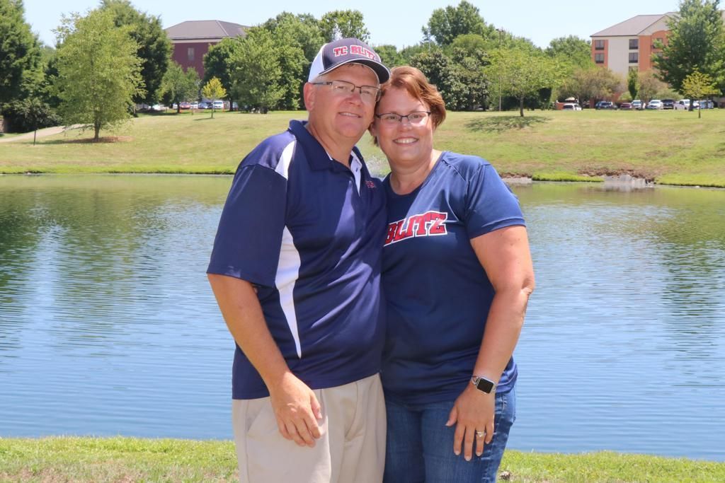 Two people smiling by a lake with trees and buildings in the background