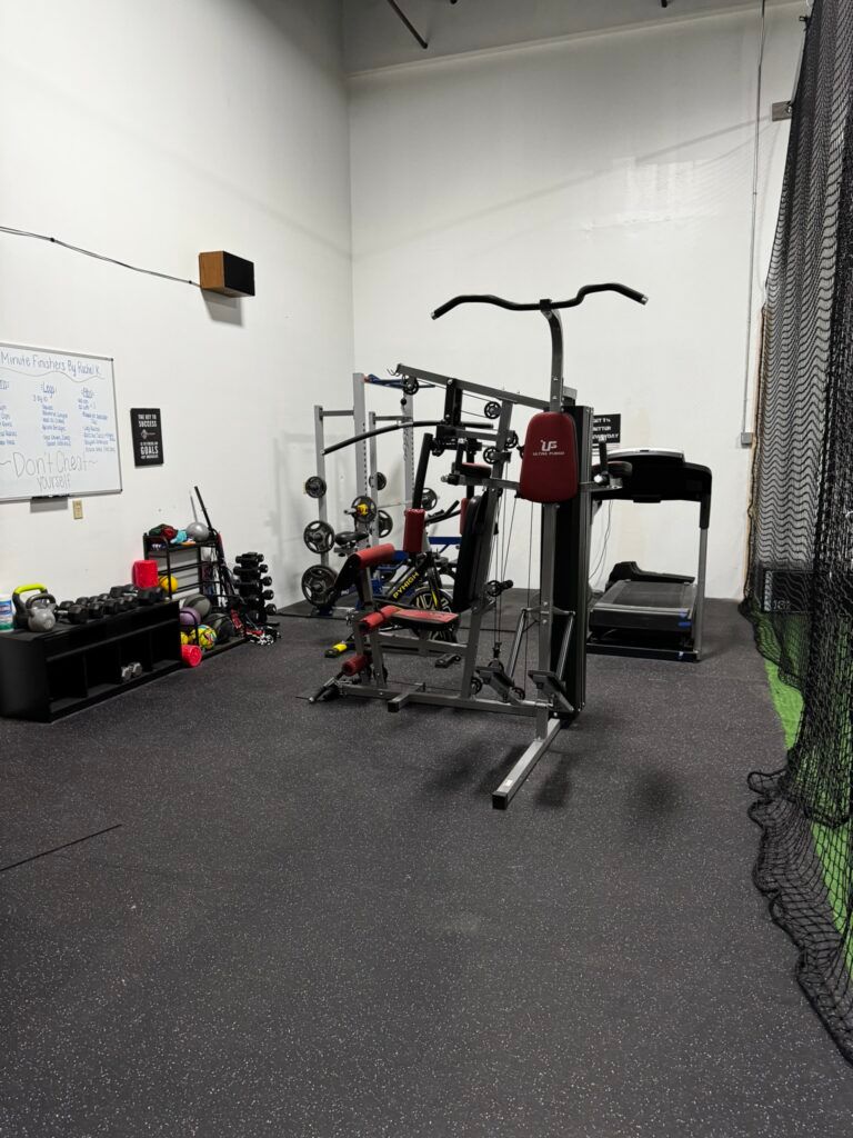Small home gym with exercise machine, weights, bench, and black rubber flooring in a white-walled room