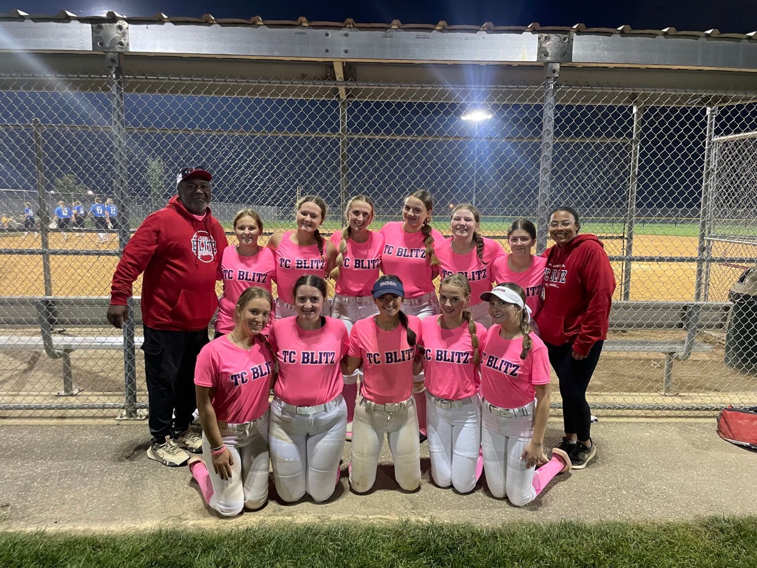 Softball team in bright pink shirts posing together on a field, with chain-link fence and dugout behind them