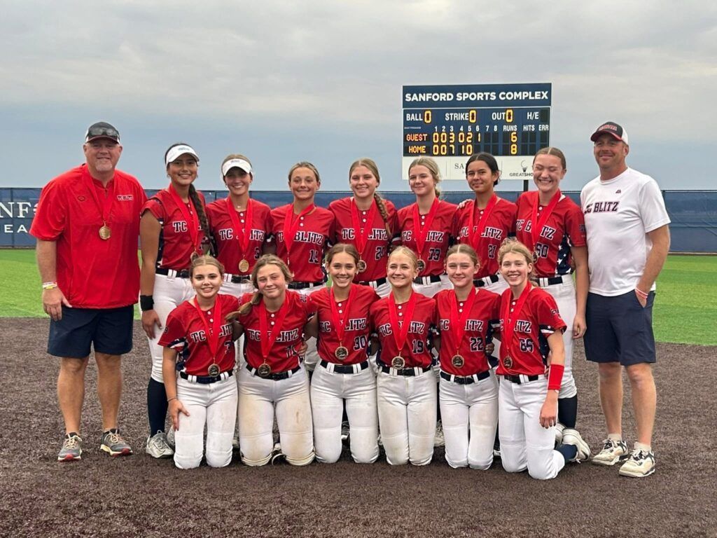 Youth softball team posing on a field with coaches; players in red jerseys and white pants.