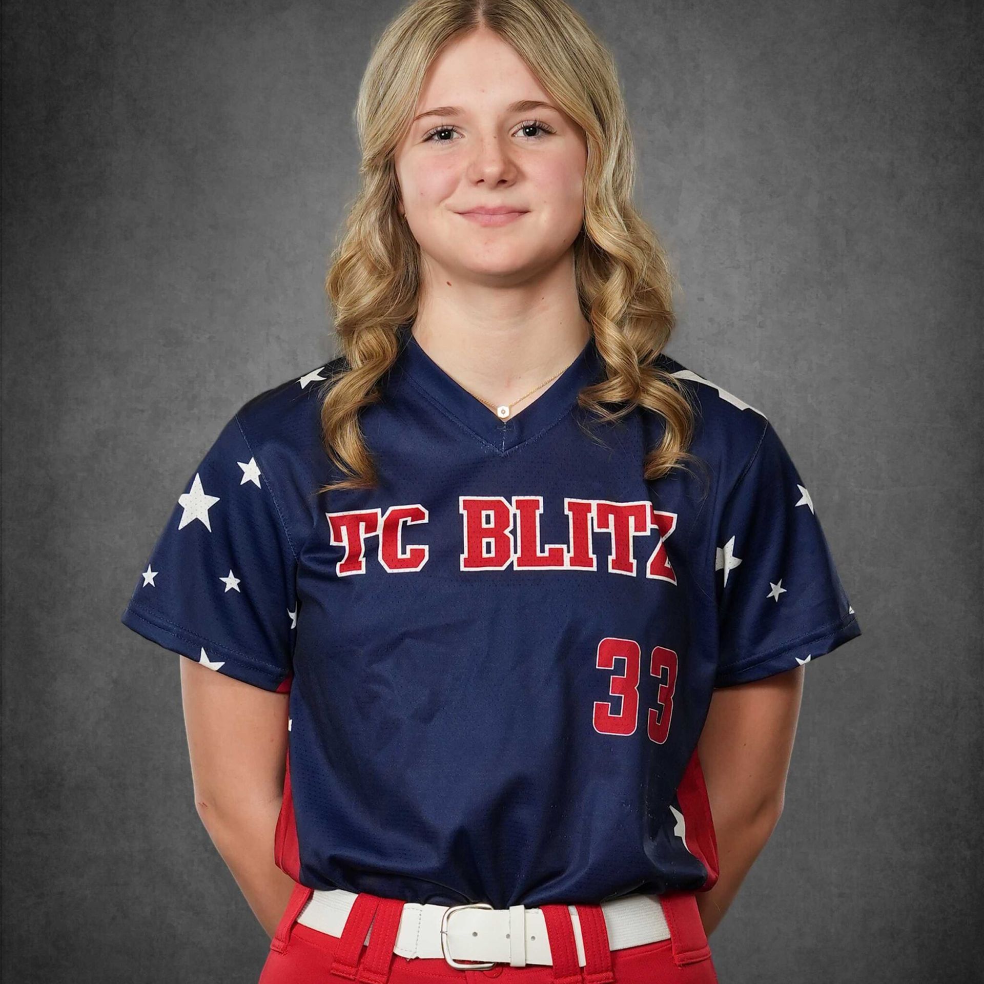 Portrait of a softball player in a navy jersey with stars, red pants, and white belt against a gray backdrop