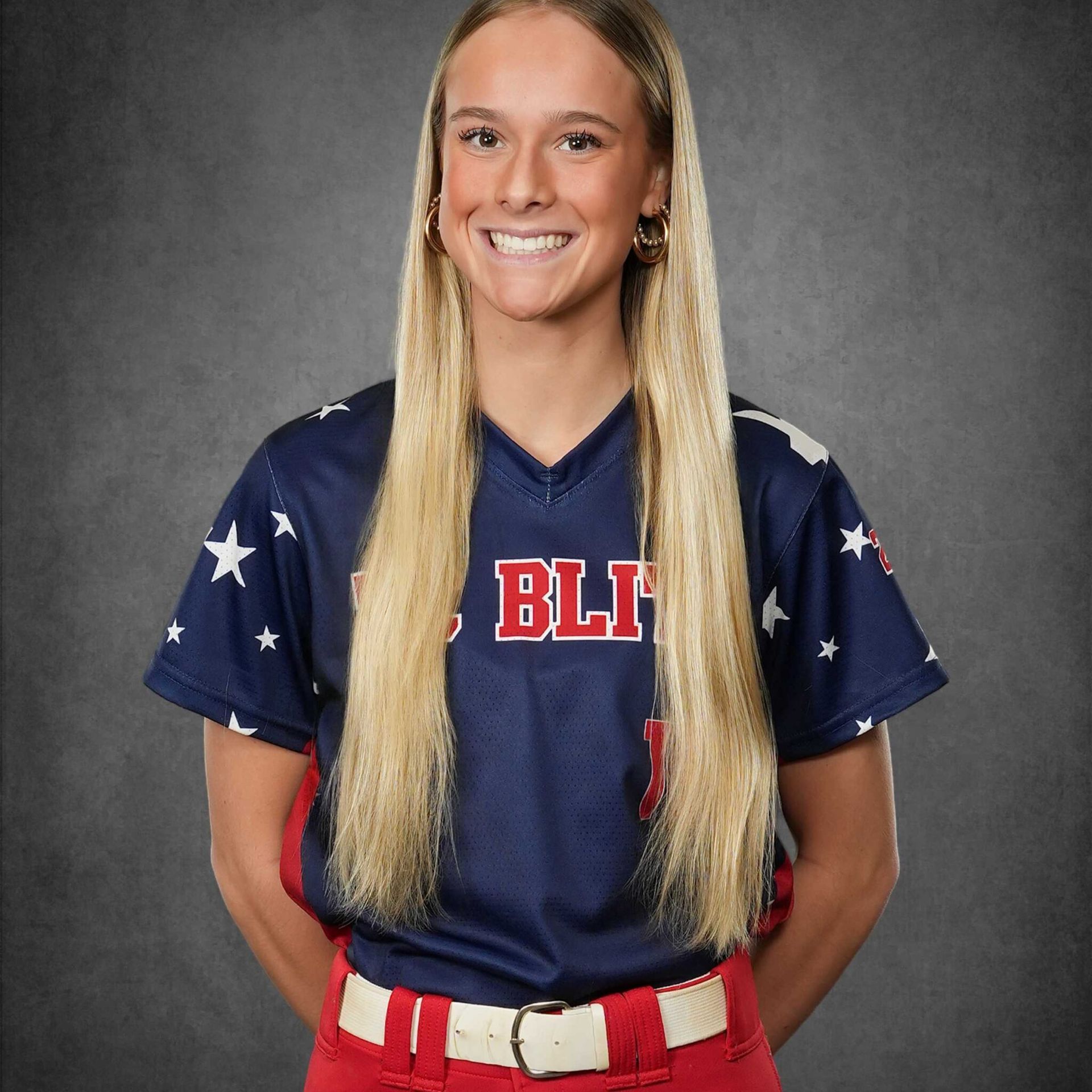 Portrait of a smiling softball player in a navy jersey with stars and red pants against a gray backdrop