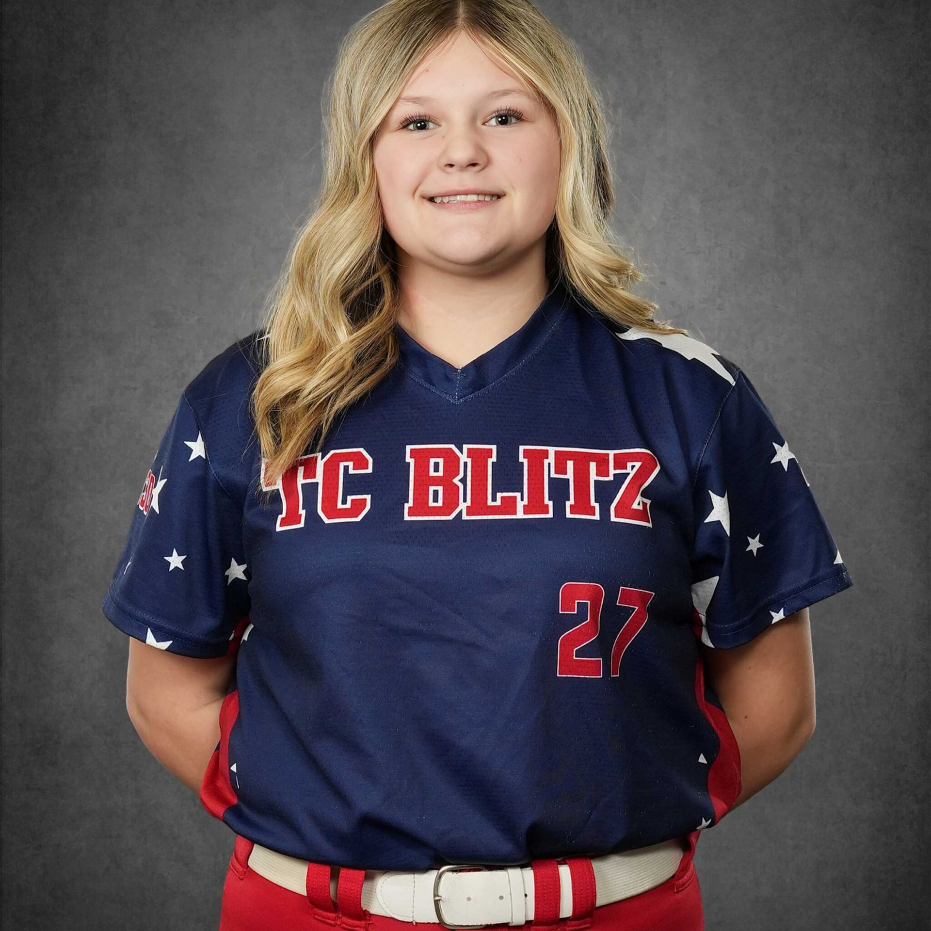 Youth softball player in navy and red uniform, standing against gray background with hands behind back.