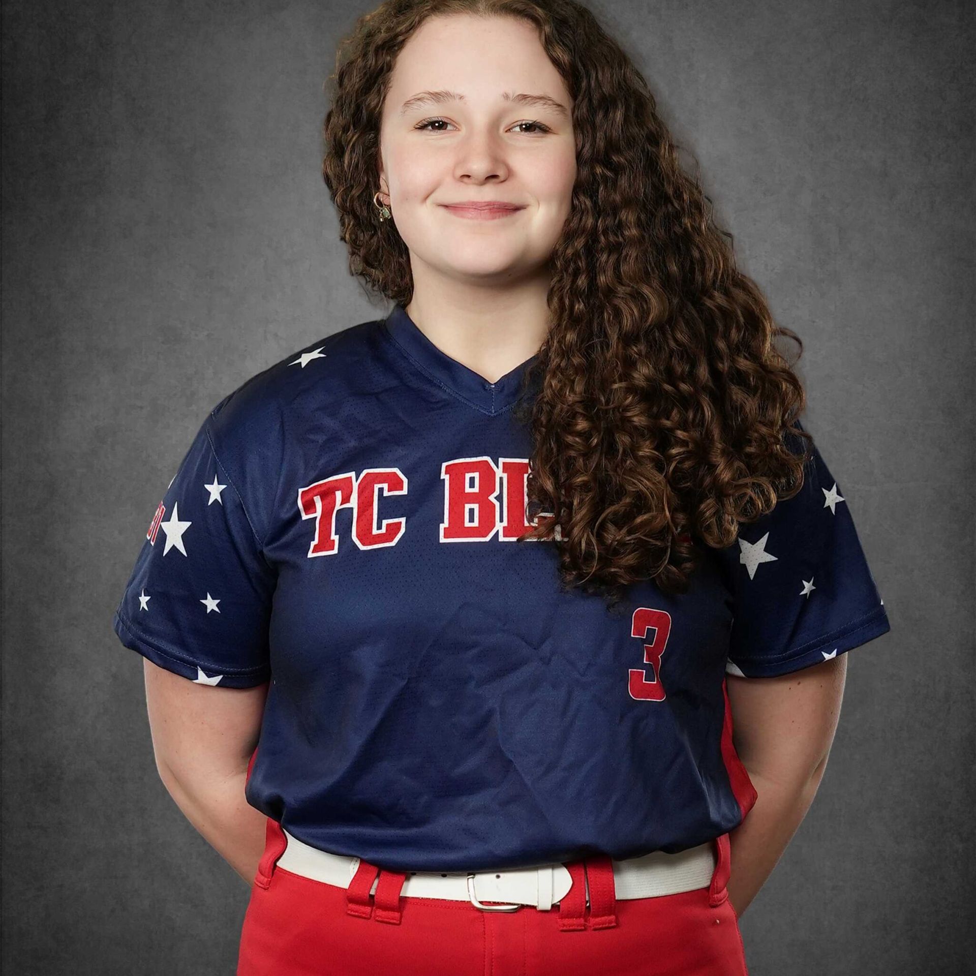 Person in a navy and red baseball uniform standing against a gray backdrop, smiling.