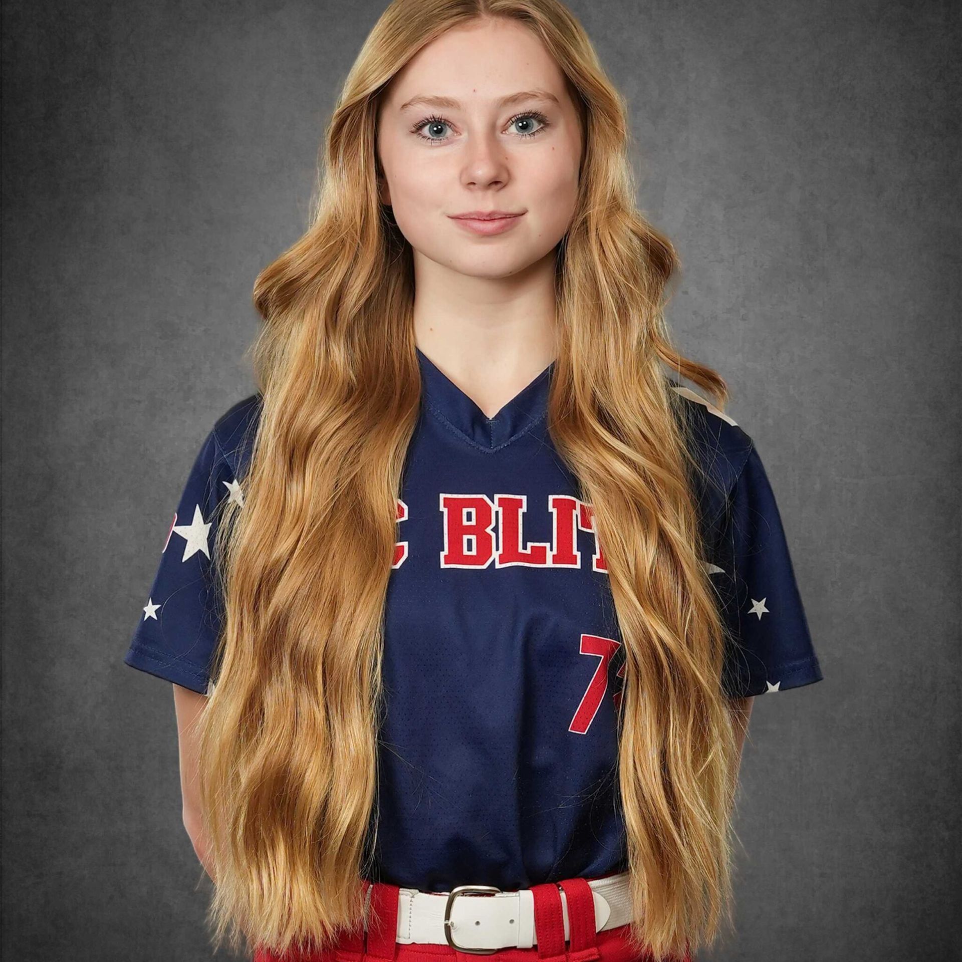Portrait of a softball player in a navy jersey with red belt, standing against a gray studio backdrop