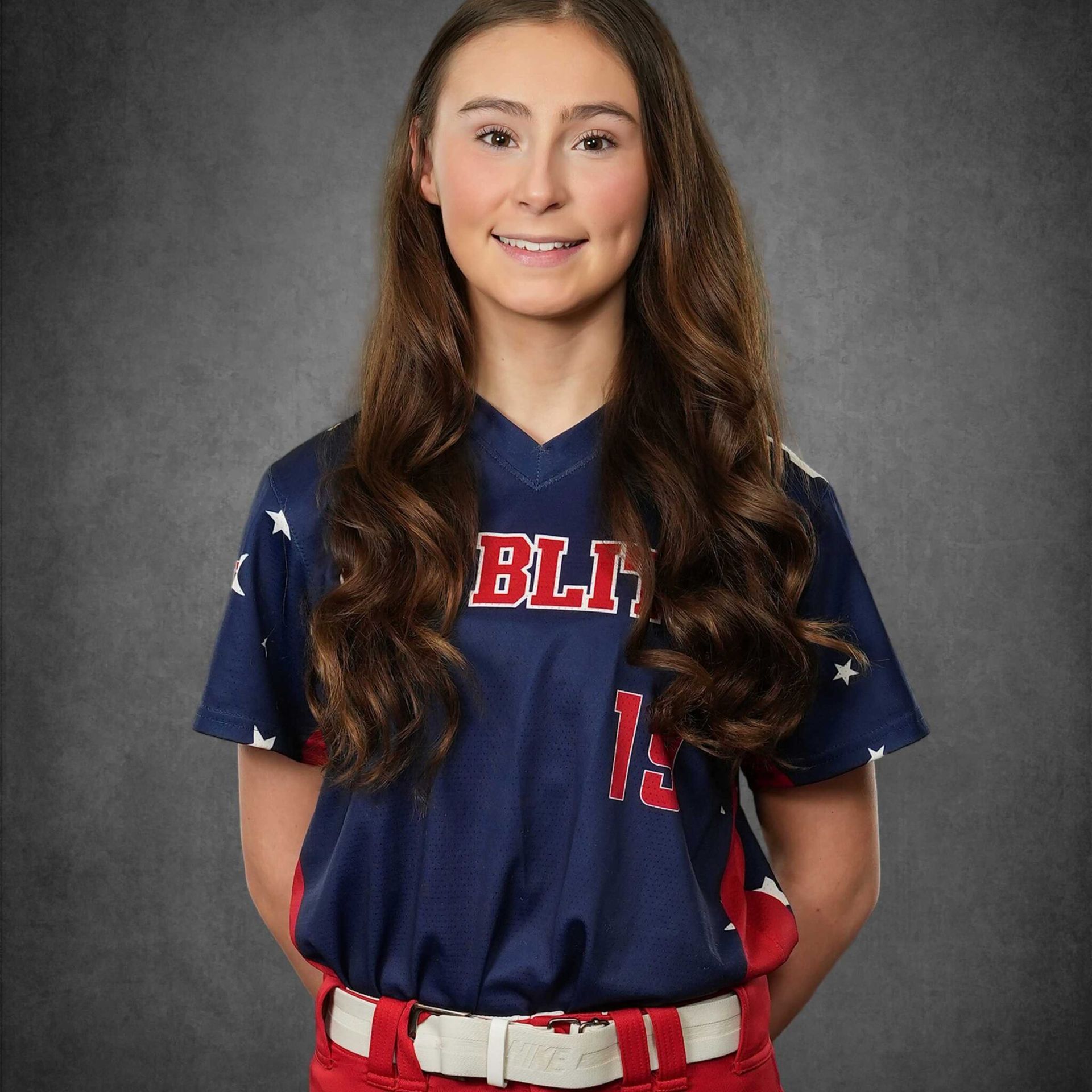 Softball player in navy jersey and red pants, smiling in front of a gray studio backdrop