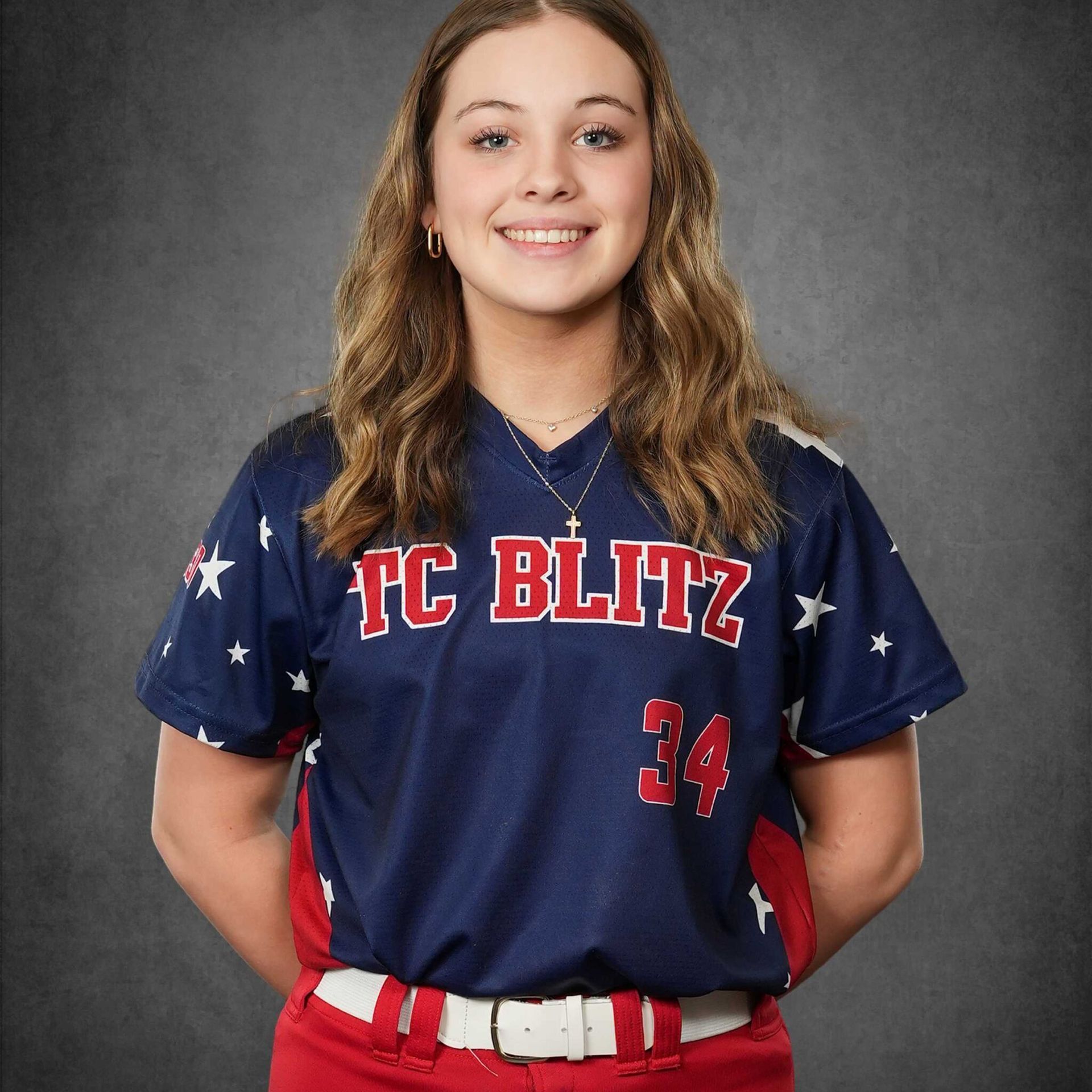 Smiling softball player in navy TC BLITZ jersey and red pants, standing against a gray backdrop
