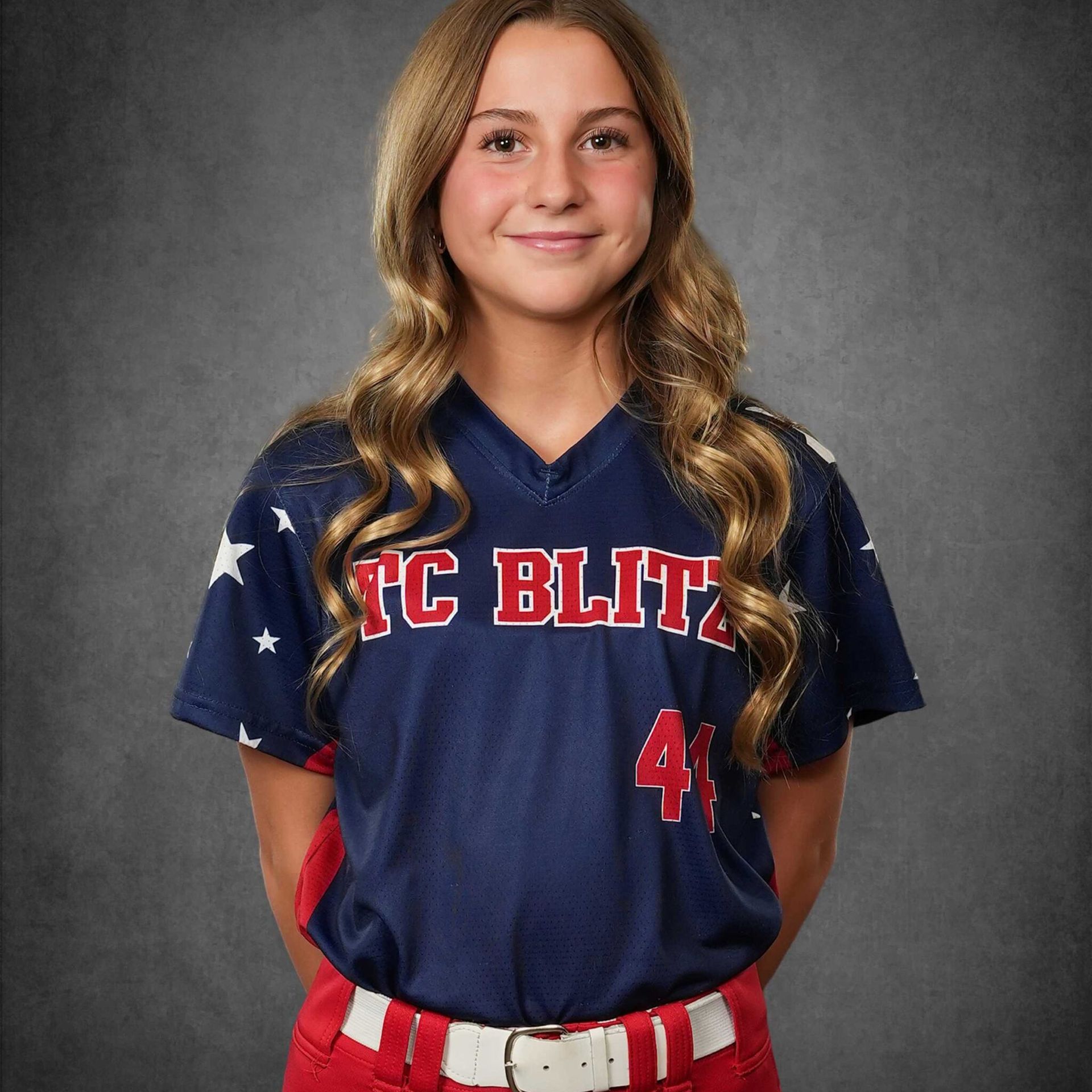 Youth softball player in navy jersey with red-and-white lettering, smiling against a gray backdrop