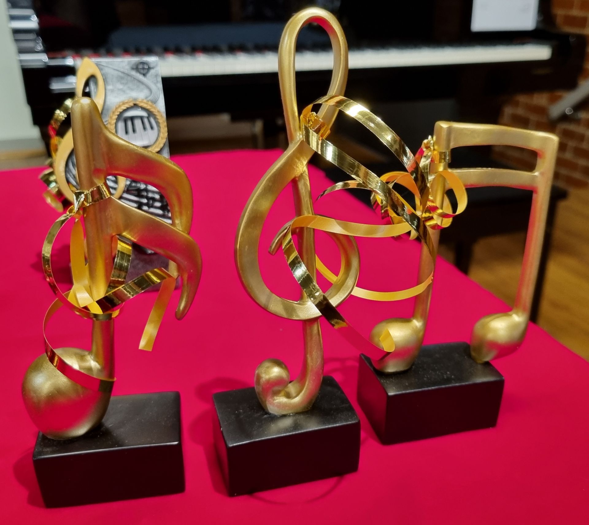 Three gold-colored musical note trophies with black bases, sitting on a red surface. A piano is in the background.