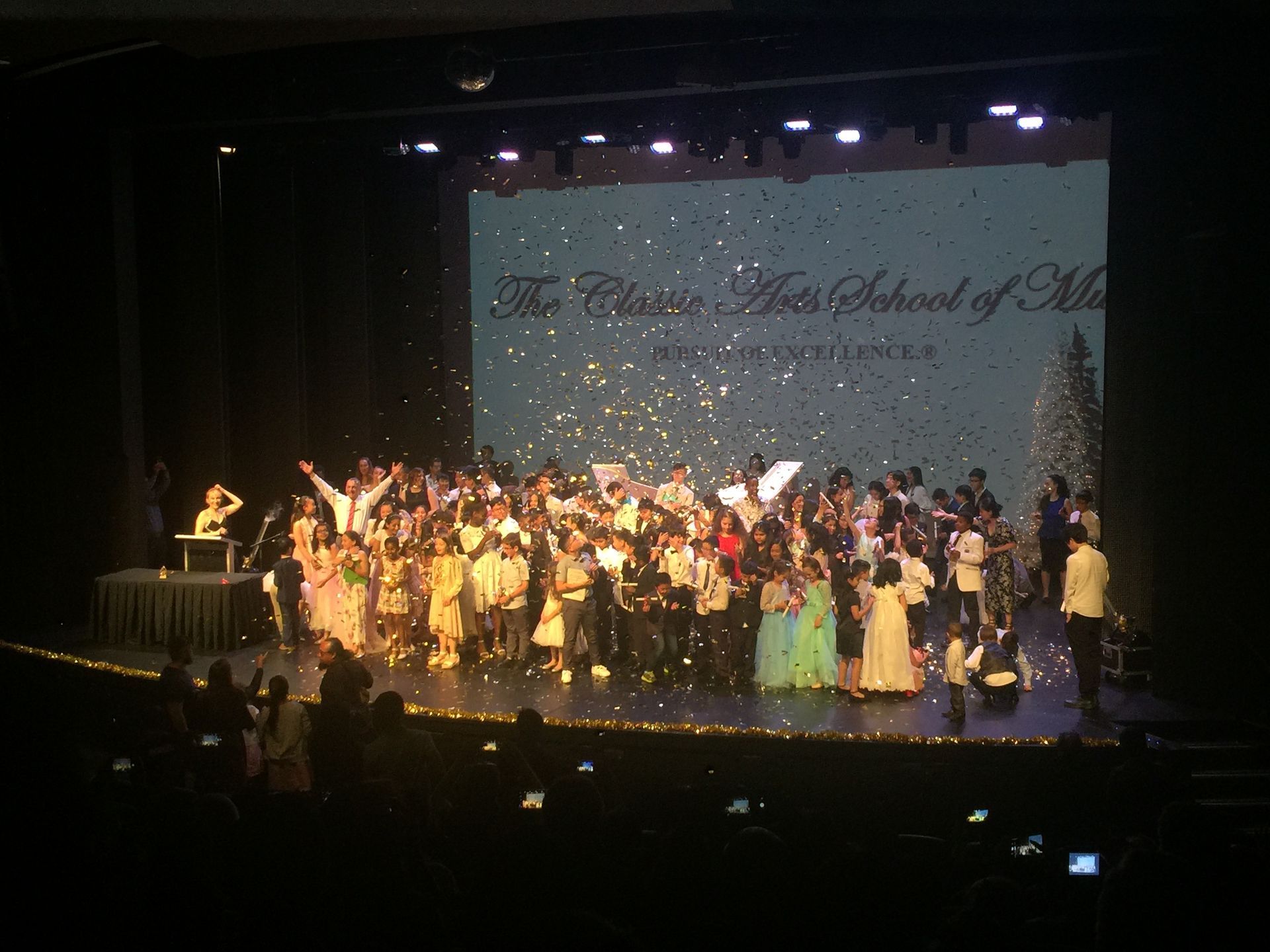 A large group of people on a stage with confetti falling. Sign reads “The Classic Arts School of Music.”