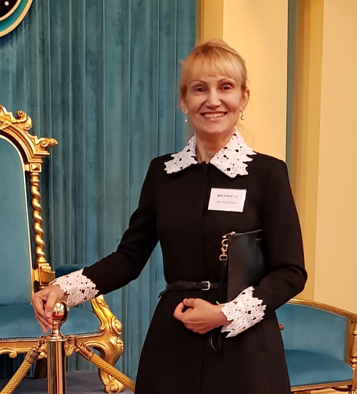 Woman in black dress with white lace trim, smiling, holding a clutch, standing by a gold and blue chair.