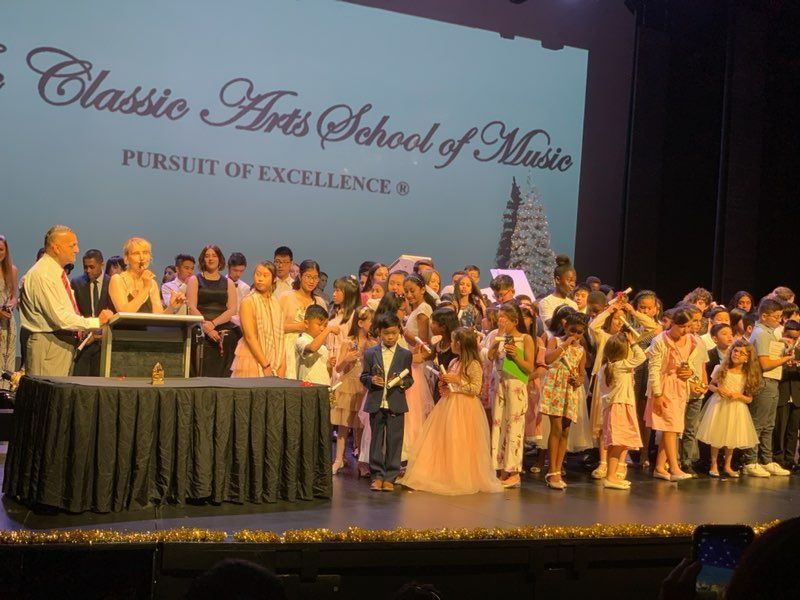Children on stage with presenters at The Classic Arts School of Music event. Large screen behind them.