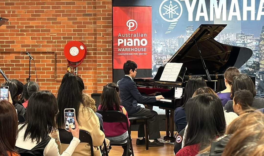 Pianist playing a grand piano for an audience; red brick wall, Yamaha sign, people with phones.