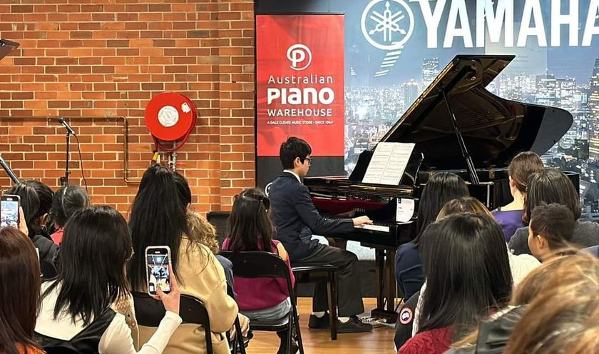 Pianist playing a grand piano for an audience; red brick wall, Yamaha sign, people with phones.