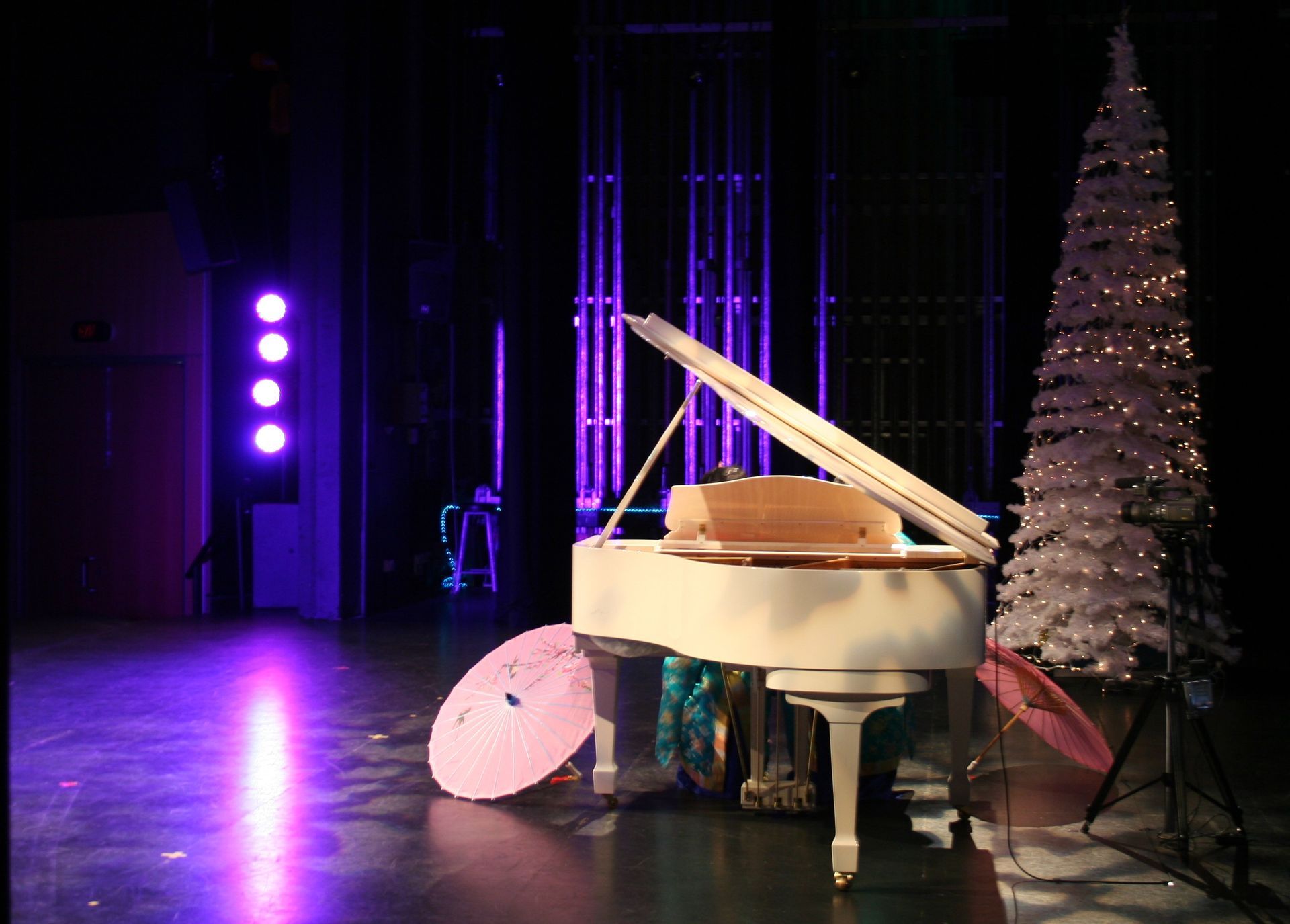 White grand piano on stage with a Christmas tree, lit with purple and white lights.