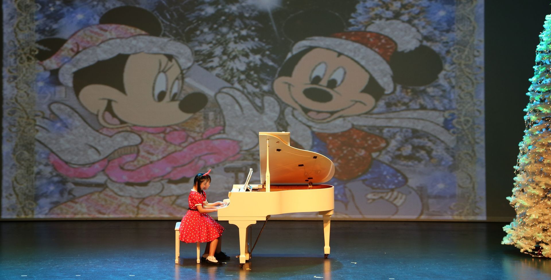 Girl playing piano on stage with Mickey and Minnie Mouse backdrop.