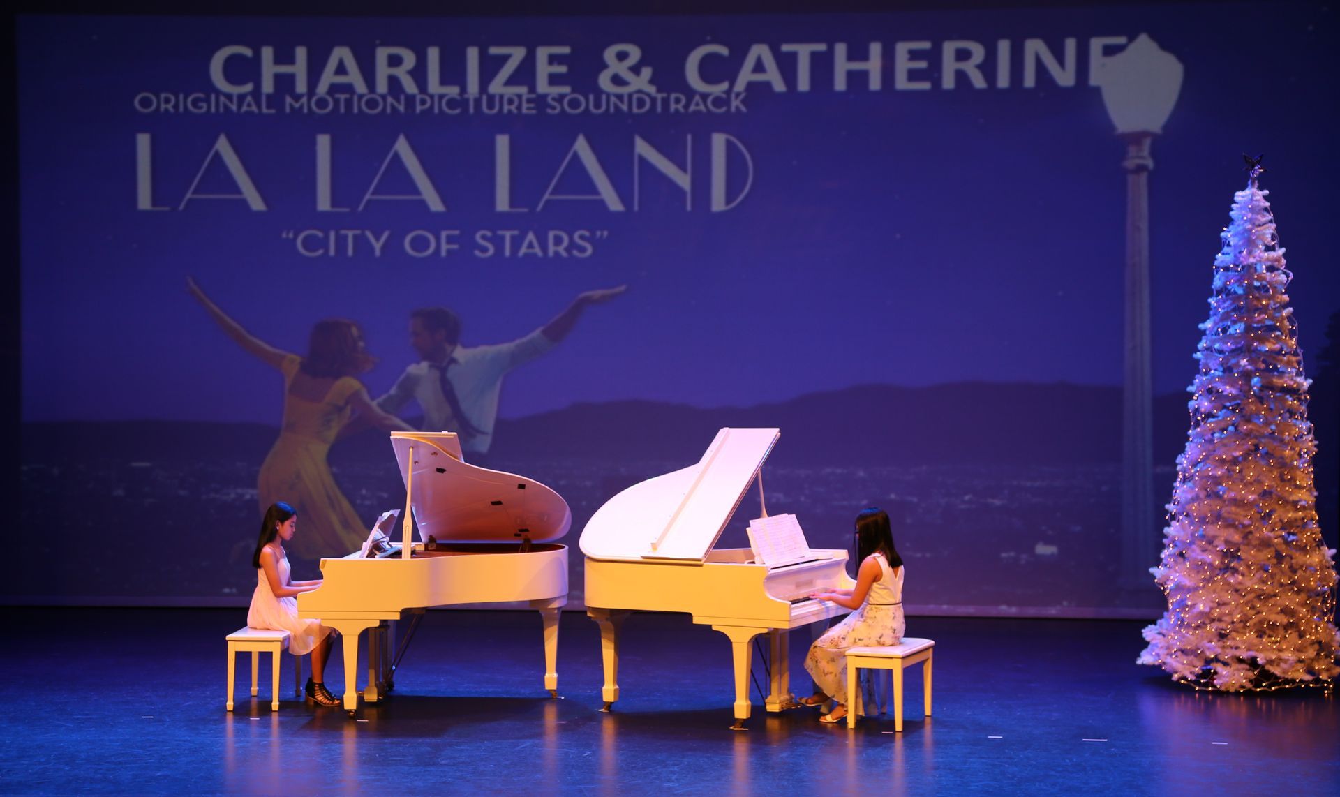 Two pianists play white grand pianos on a stage with a 