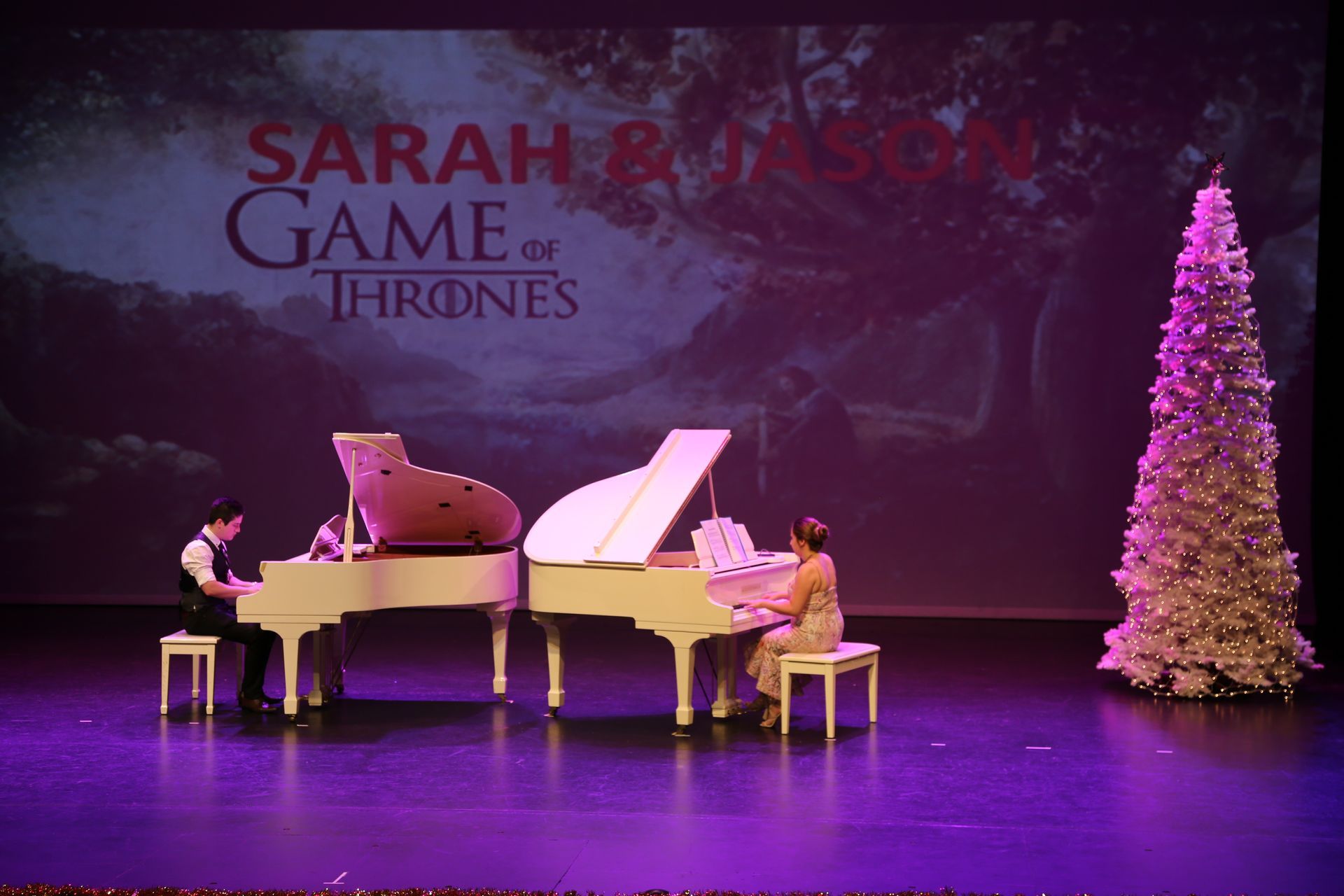 Two pianists play white pianos on a stage with a Christmas tree and a Game of Thrones backdrop.