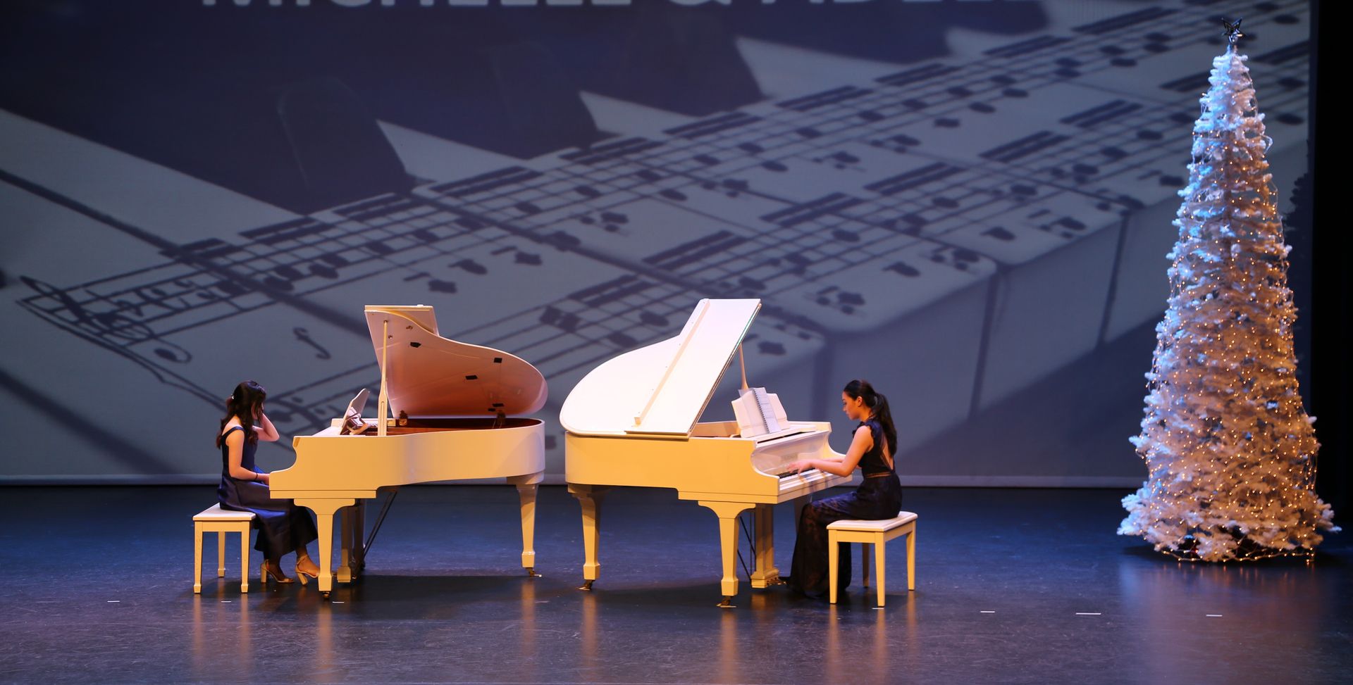 Two pianists perform on stage with white and cream pianos, Christmas tree backdrop.