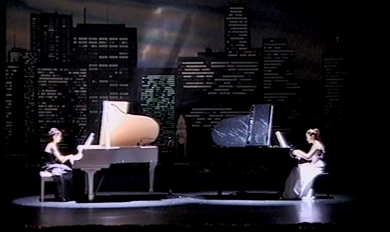 Two pianists playing on stage with a city skyline backdrop; a white and a black piano.