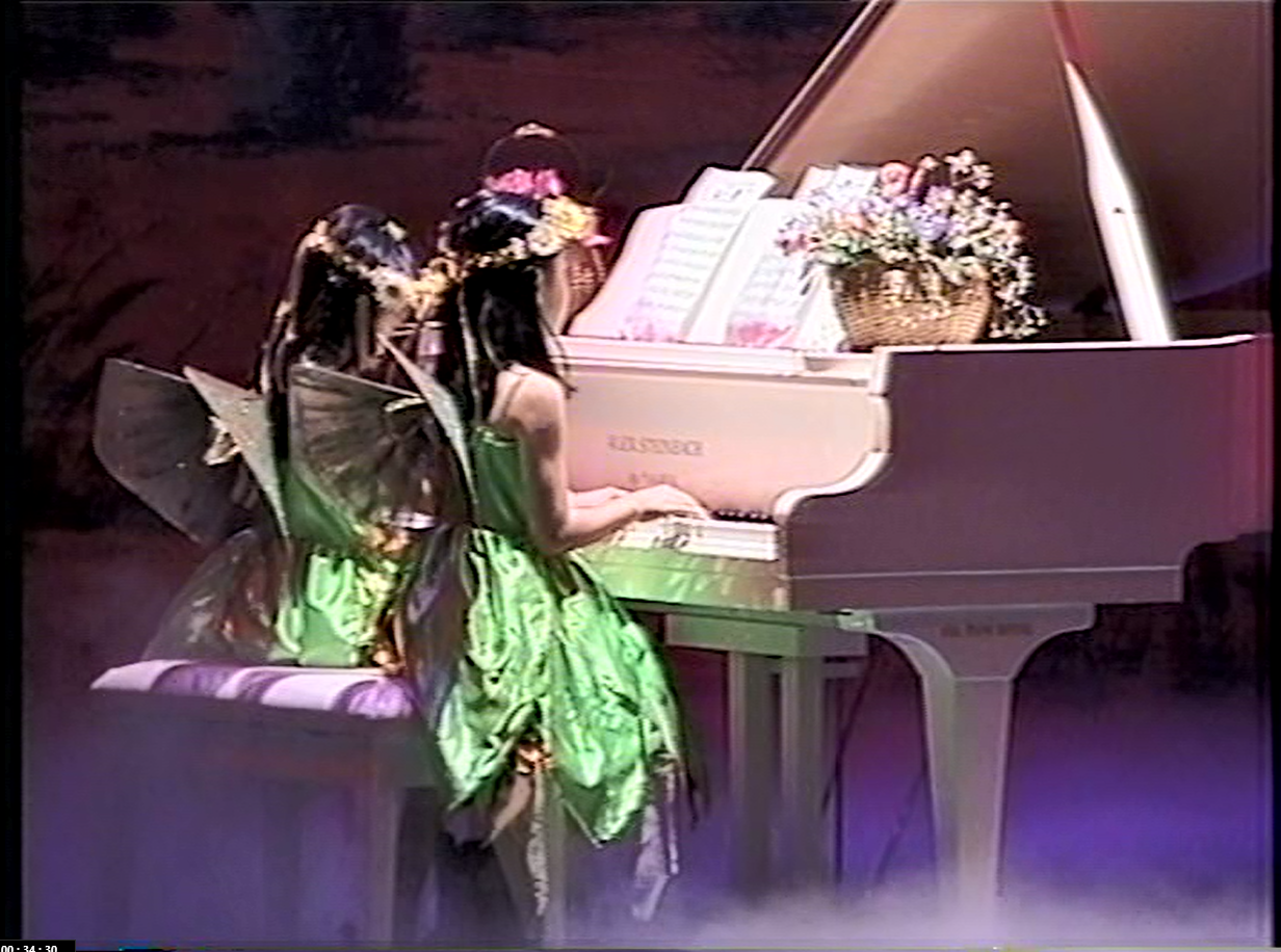 Two girls in fairy costumes playing a white piano, flowers on top, stage setting.