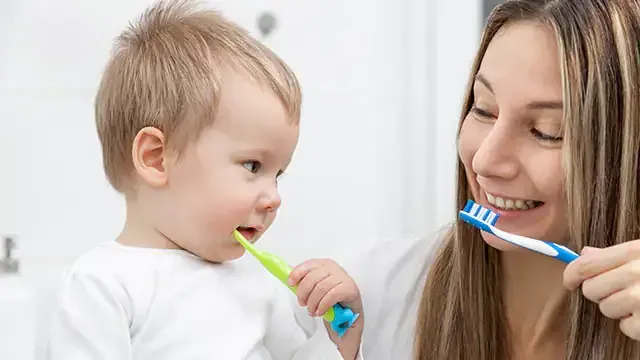 Woman and child brushing teeth together in a bathroom, smiling. They each hold a toothbrush.