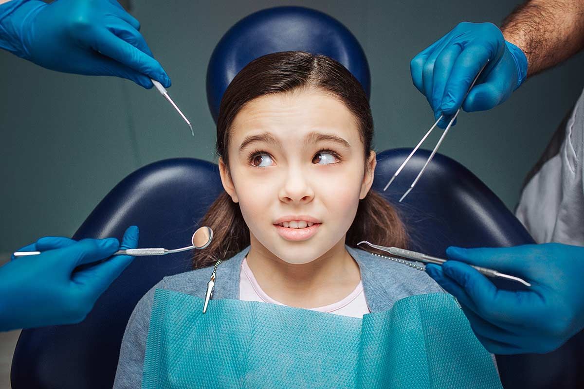 Girl in dental chair surrounded by gloved hands holding dental tools, looking anxious.