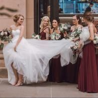Bride and bridesmaids in burgundy dresses lifting the bride’s flowing white gown outdoors