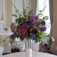 Tall floral centerpiece with purple, pink, and green flowers in a white vase on a table in an elegant room