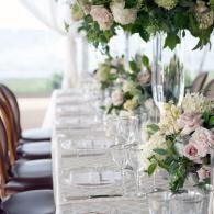 Elegant wedding reception table with white floral centerpieces, glassware, and wooden chairs by a bright window