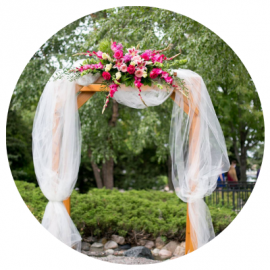 Outdoor wedding arch with white drapes, pink floral arrangement, and greenery in a garden setting