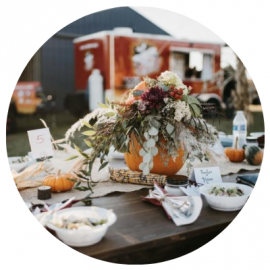 Outdoor autumn table with pumpkin centerpiece, flowers, and bowls of food at a rustic gathering
