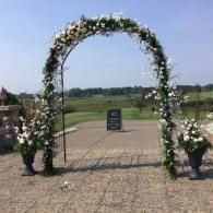 Flower-covered wedding arch on a paved path with open fields in the background