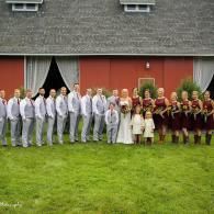 Large group posing on grass in front of a red barn, with draped white curtains and formal outfits
