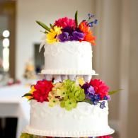 Two-tier white wedding cake topped and wrapped with bright multicolored flowers