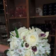 White and purple floral bouquet in a vase, with glassware and dark shelves in the background