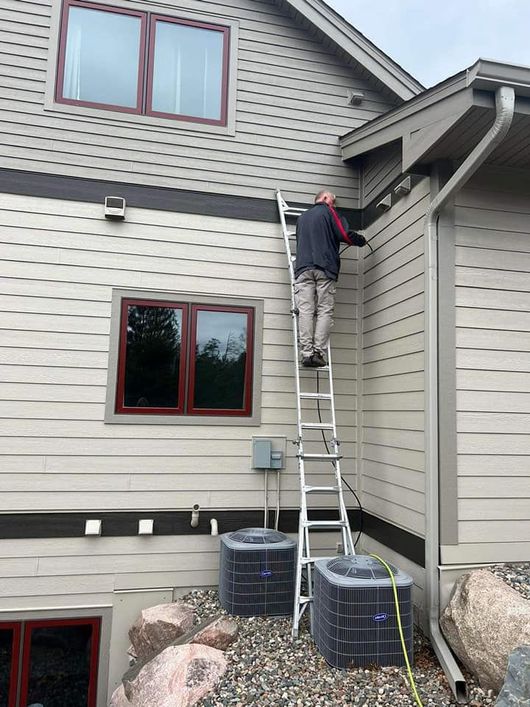 An specialist from a air duct cleaning company is standing on a ladder on the side of a house cleaning an air duct.