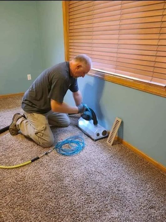 A air duct cleaning professional is kneeling on the floor in a room providing central air unit cleaning services.