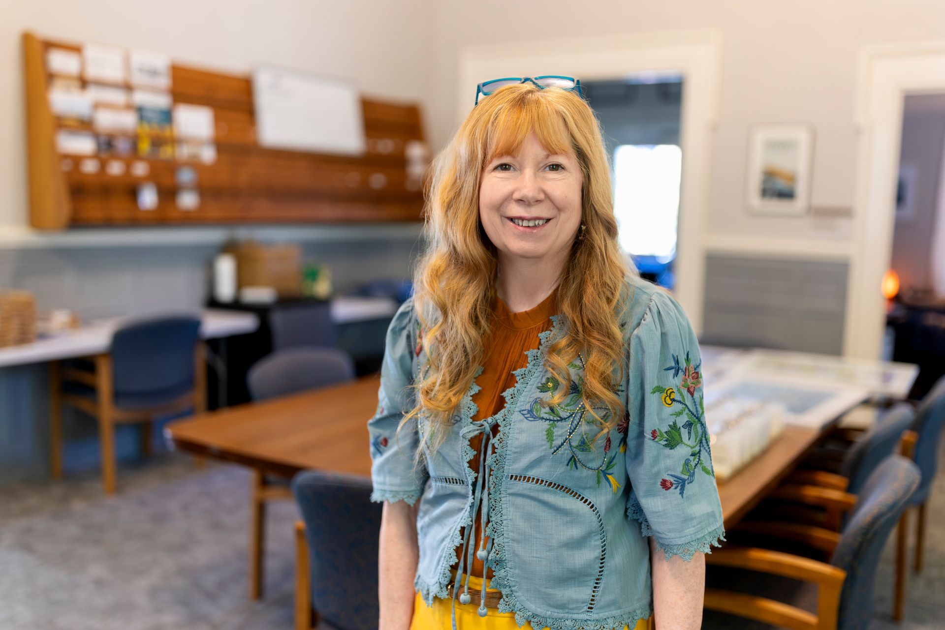 Woman with blonde hair smiles, wearing a blue floral top and tan cardigan indoors, blurred background.
