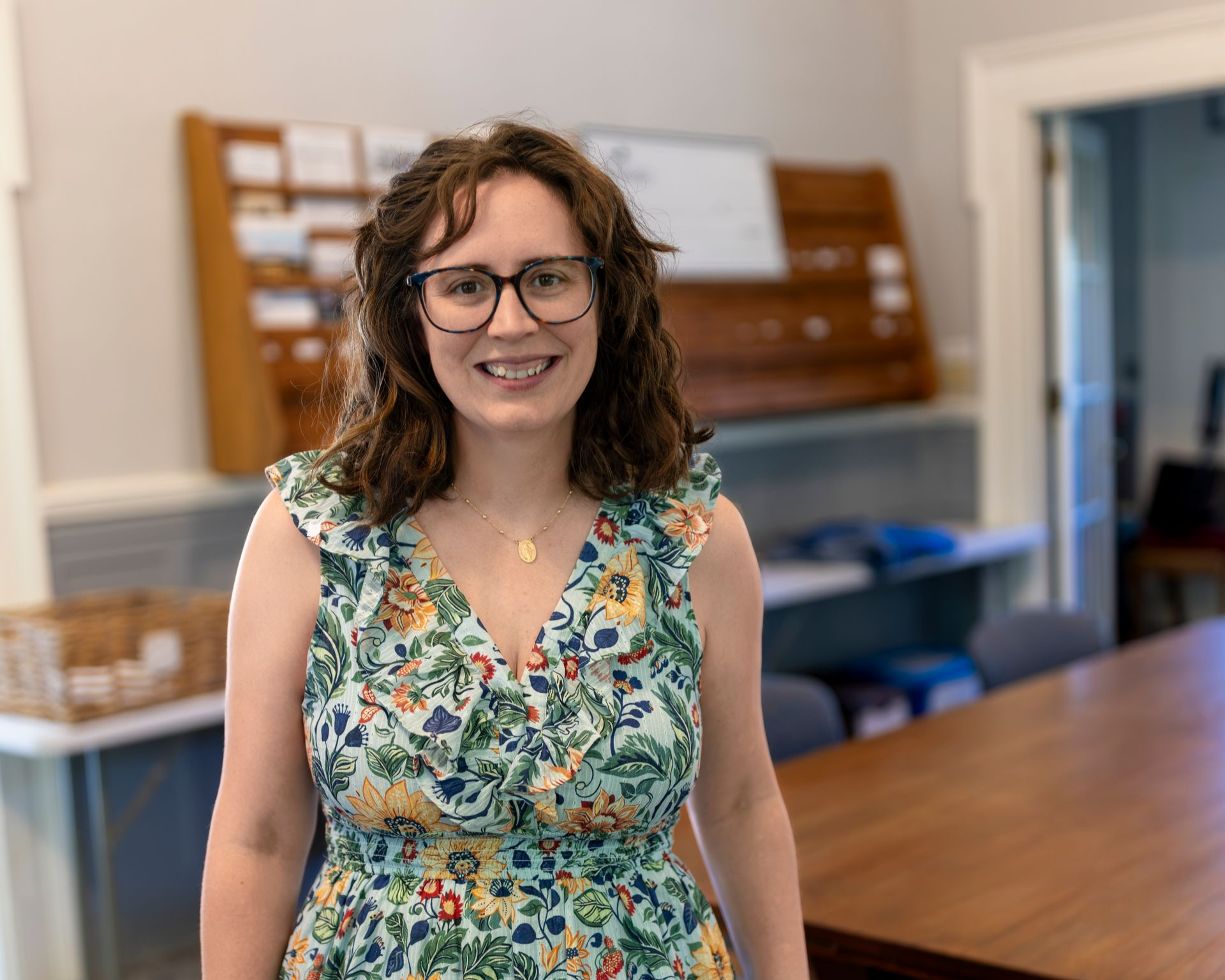 Smiling woman in glasses and floral dress standing in a bright room with a wooden cabinet and table behind her