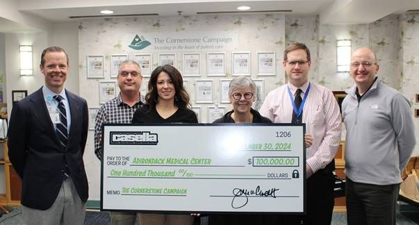 Six people hold a large check. The check is green and white. They're in a modern room with artwork on the wall.