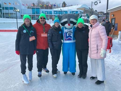 Group of six people and a raccoon mascot on an ice rink, all wearing green hats.