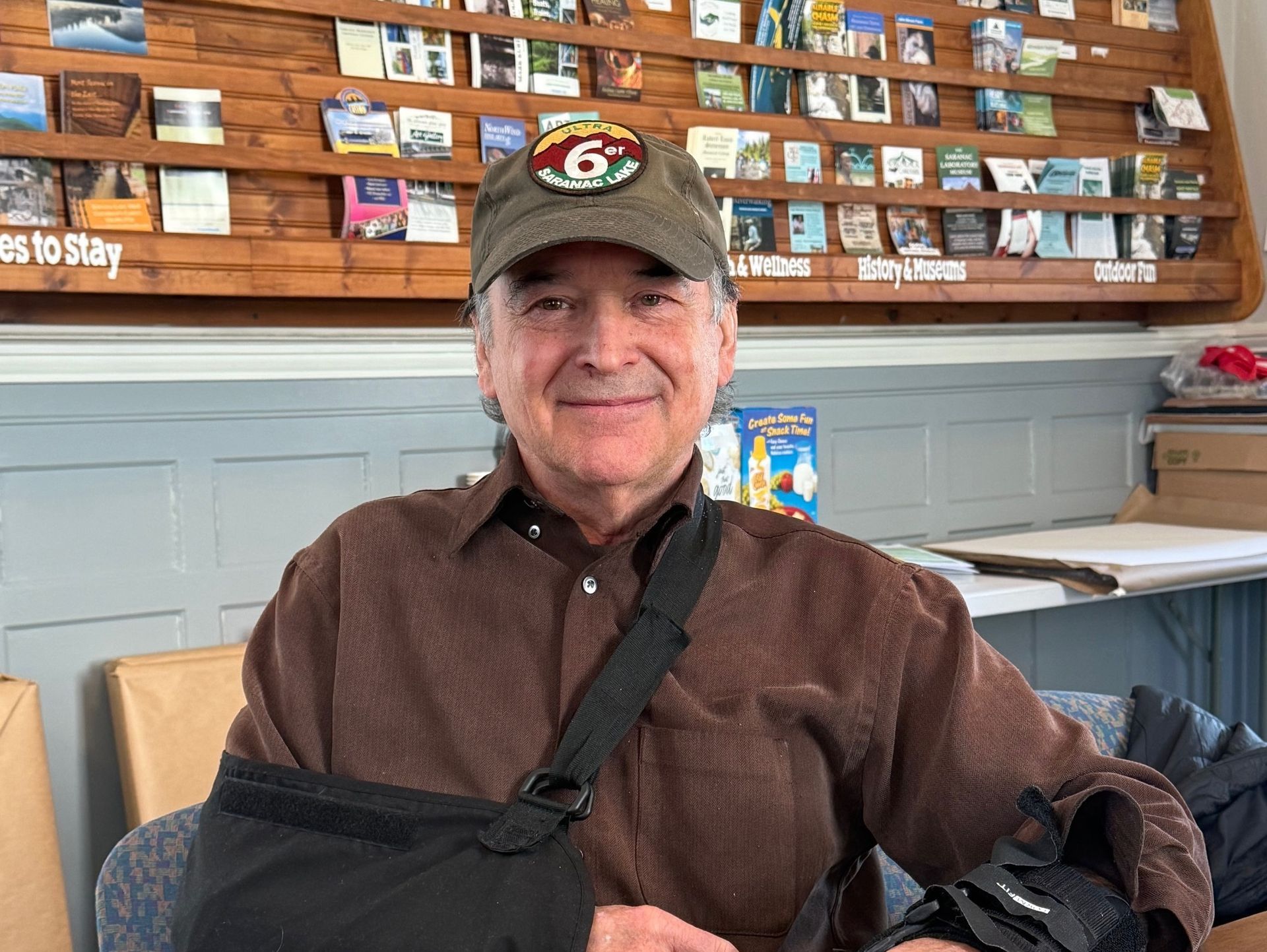 Man with a baseball cap and arm in a sling smiling, indoors in front of a wooden shelf with books.