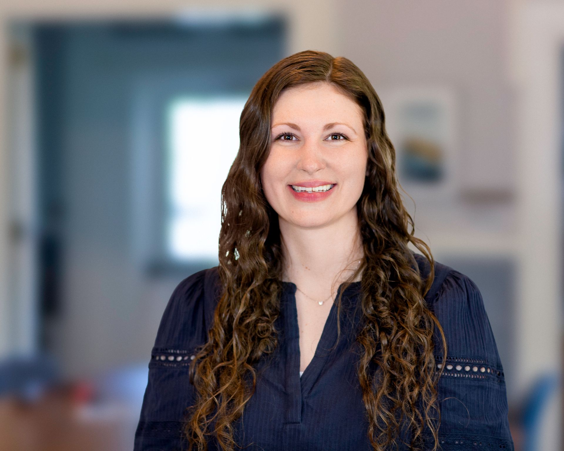 Woman with long, curly brown hair smiling at the camera, wearing a navy blue top, standing in a home setting.