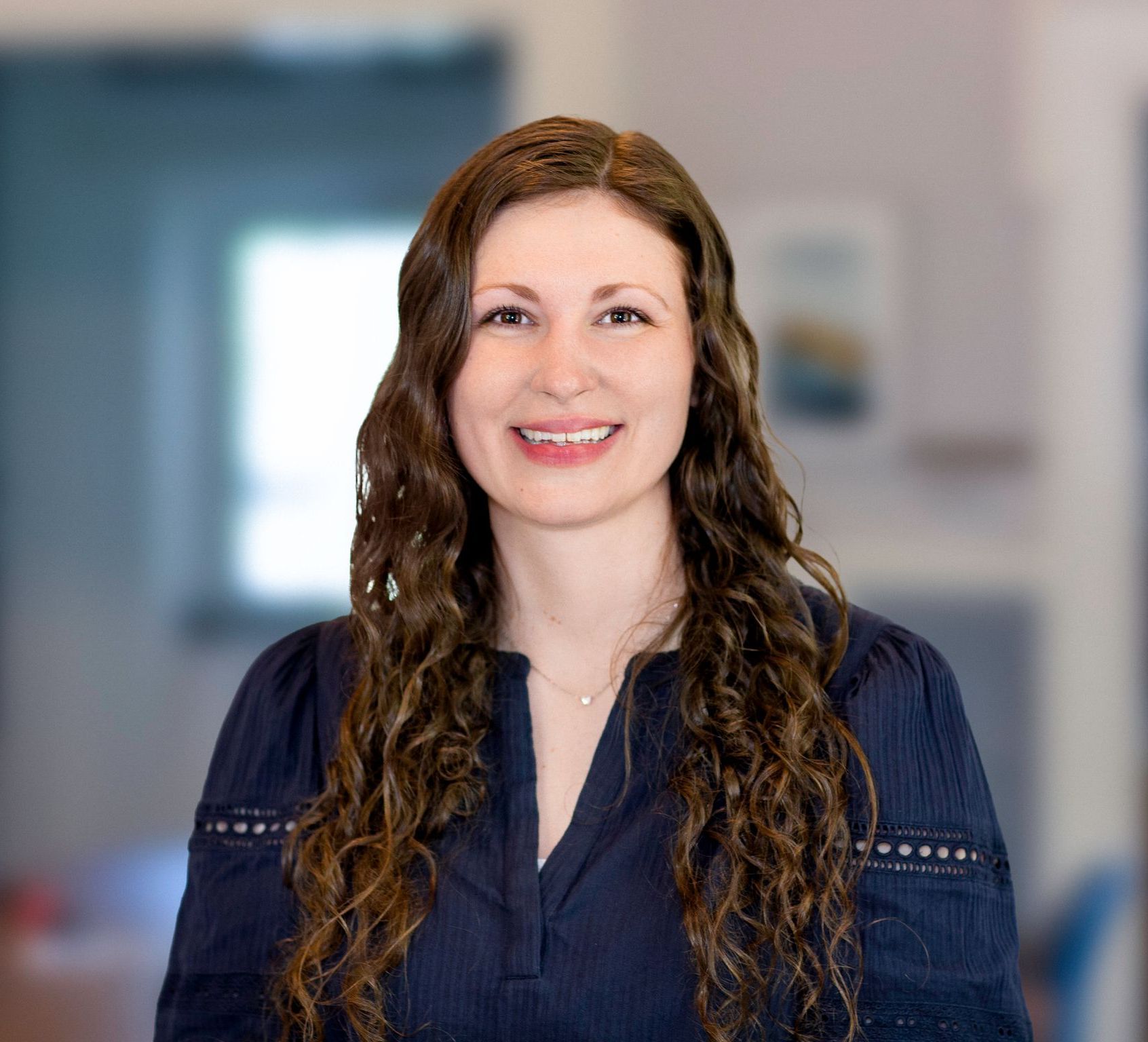 Woman with long, wavy brown hair smiles, wearing a blue shirt, indoors with a blurred background.