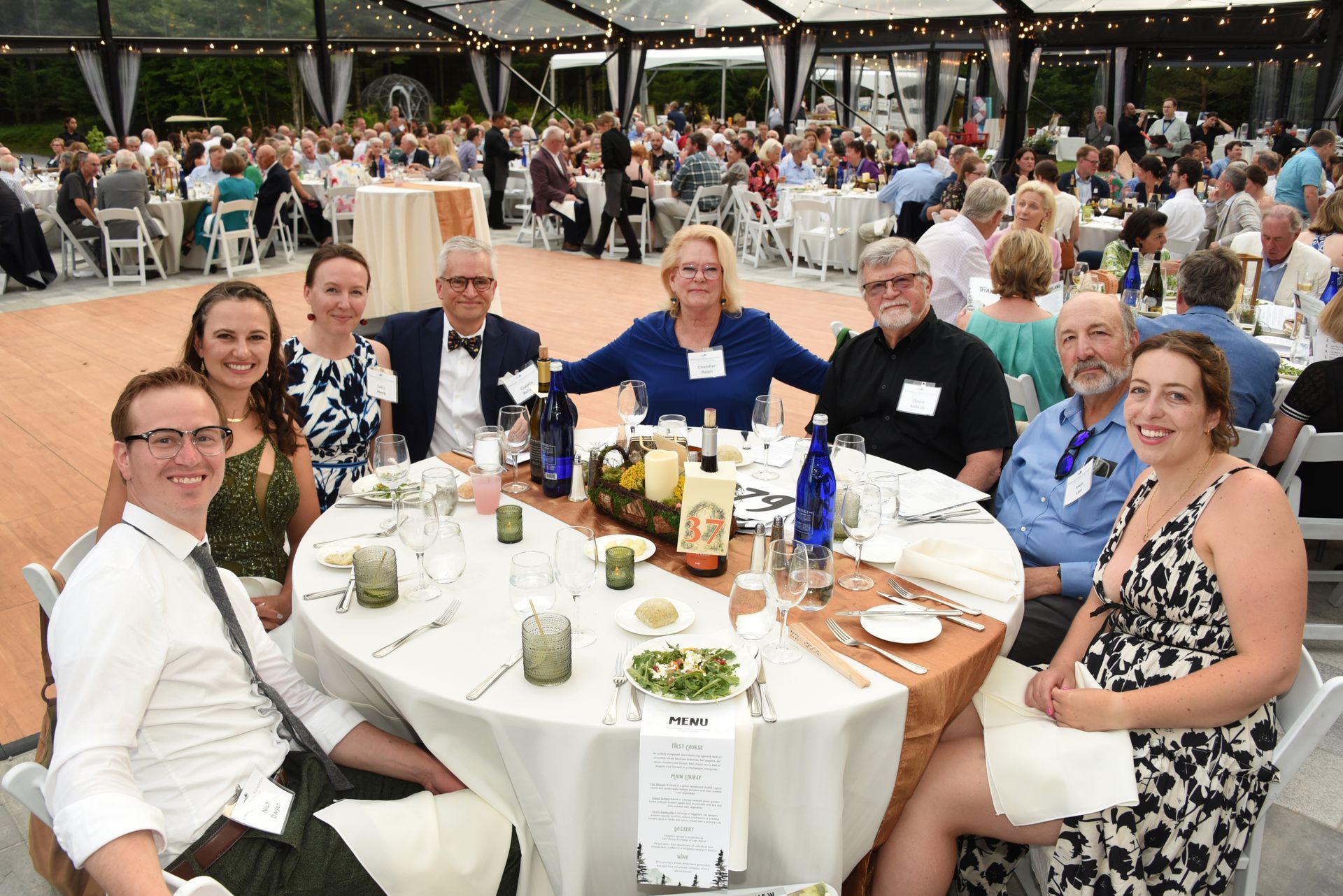 Group of people seated at a round table at an outdoor event. They are smiling and interacting with each other.