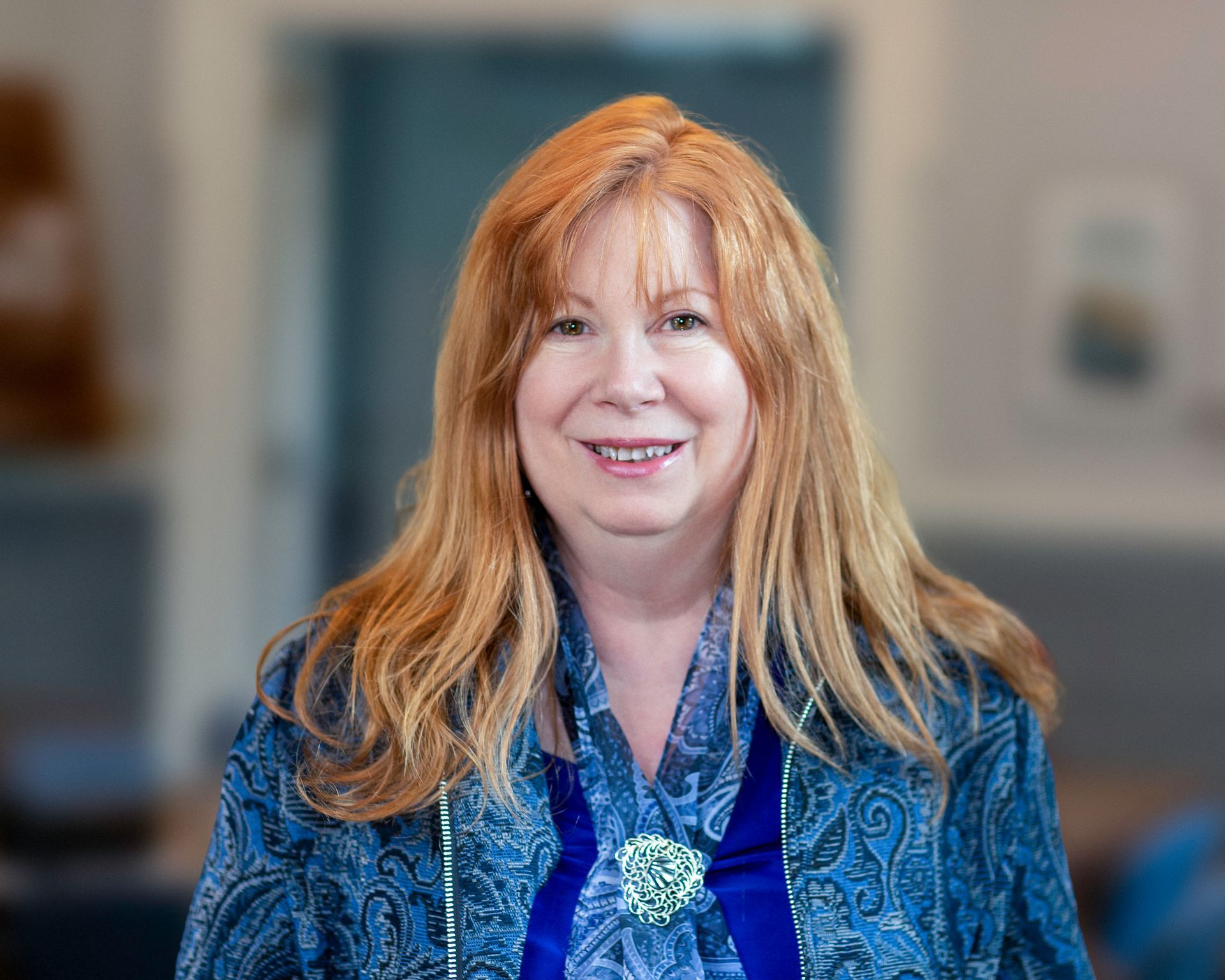 Woman with blonde hair smiles, wearing a blue floral top and tan cardigan indoors, blurred background.