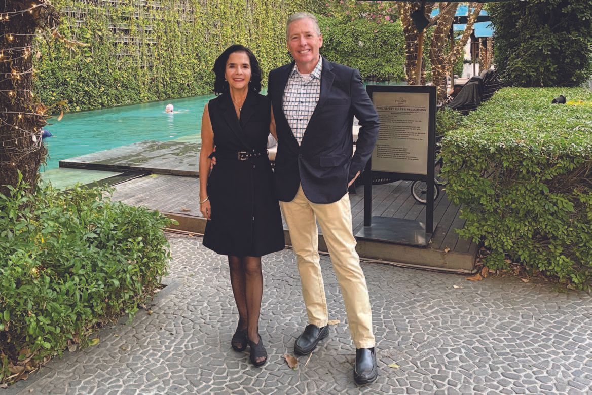 A woman and a man pose outdoors. The woman wears a black dress, and the man wears a blazer and khakis.