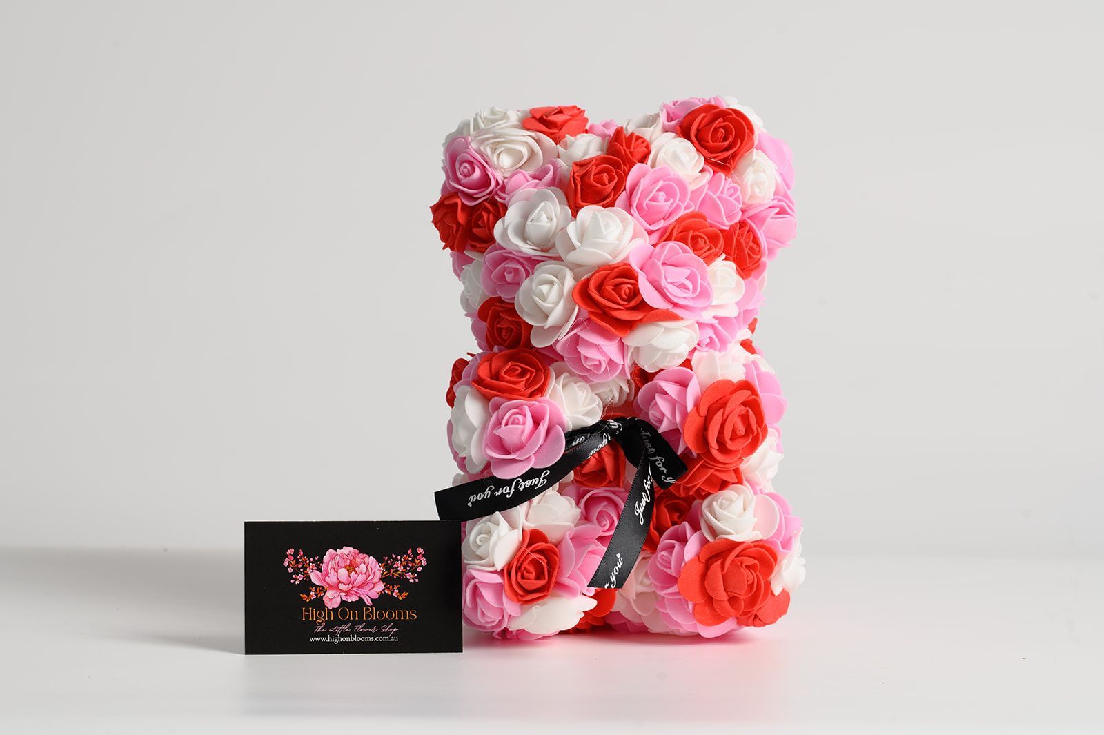 Rose bear made of pink, red, and white flowers, with a black ribbon. A black card is on the left.